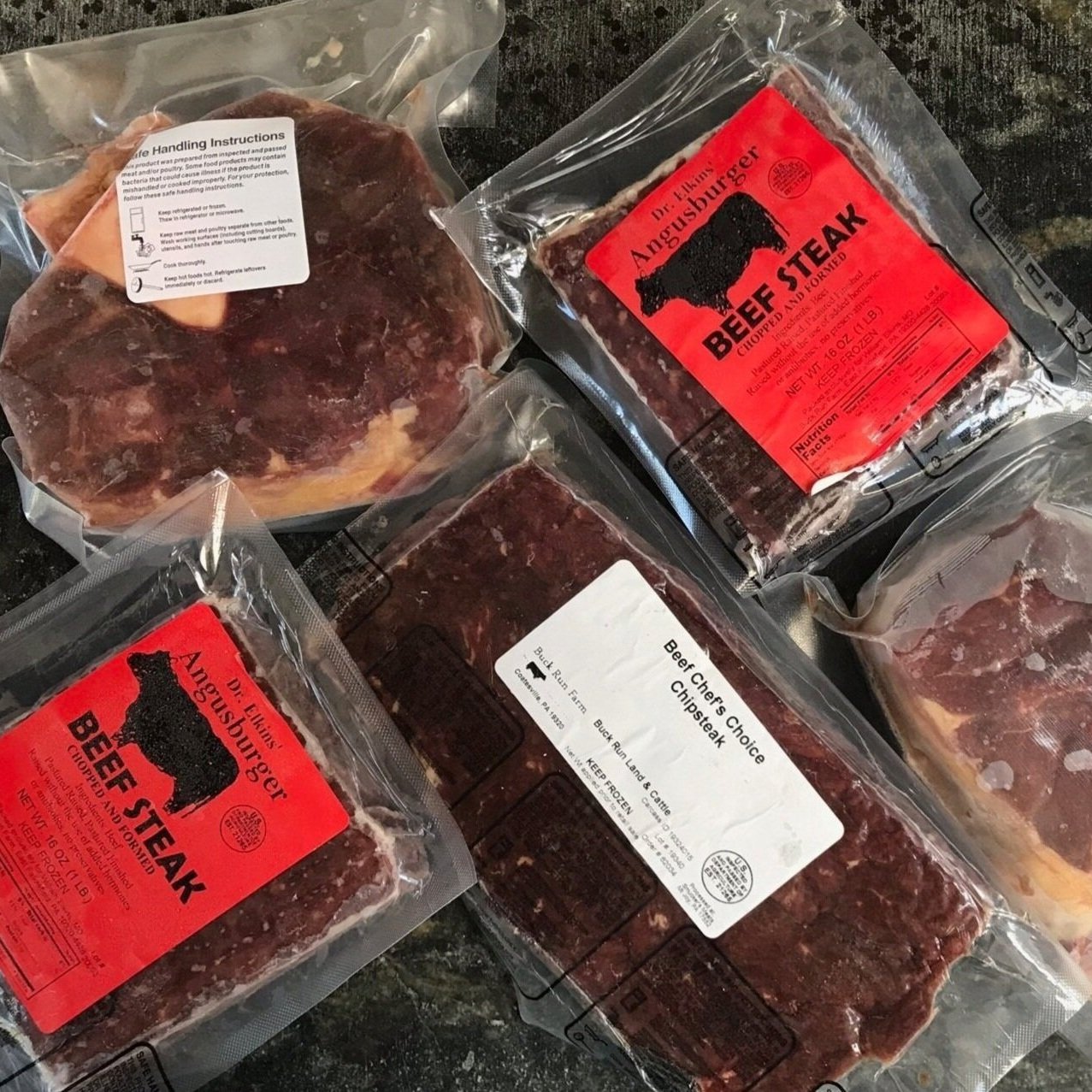 Assorted vacuum-sealed packages of Angus beef products, including beef steaks and hamburgers, featuring clear labels and handling instructions, arranged on a dark countertop.