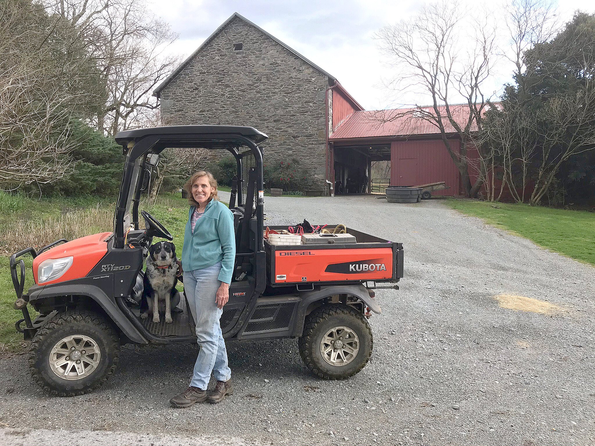 Woman standing next to a Kubota utility vehicle with a dog, on a gravel path in front of a rustic barn, surrounded by trees and farmland.